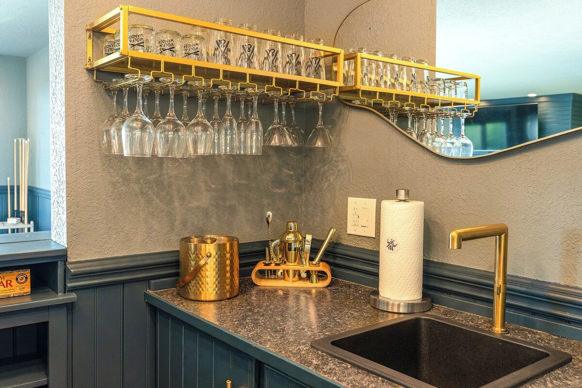 Un coin bar avec une étagère où sont rangés des verres à vin, un évier doté d’un robinet doré et un comptoir de bar agrémenté d’un objet décoratif.