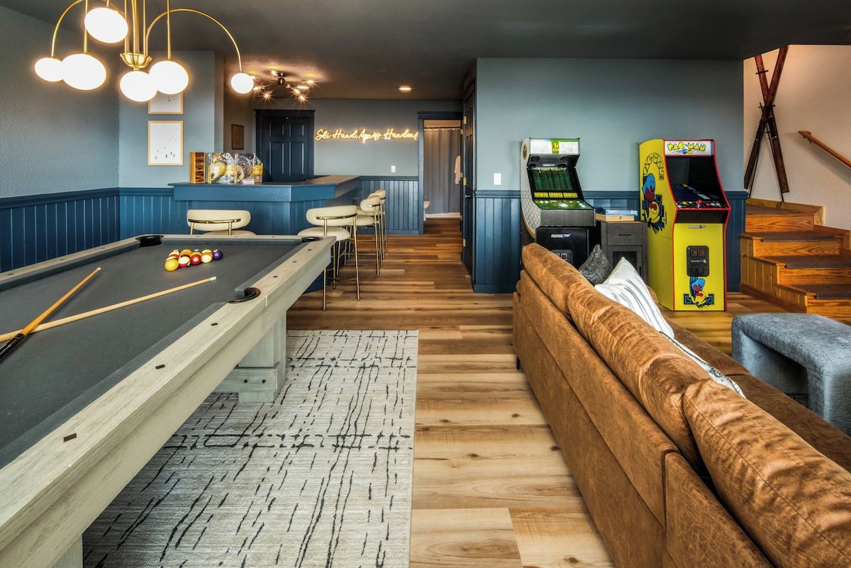Ein Billardtisch mit Kugeln, eine Billardkeule, eine Couch und ein Neon-Leuchtschild in einem Raum mit Holzboden und blauen Wänden.