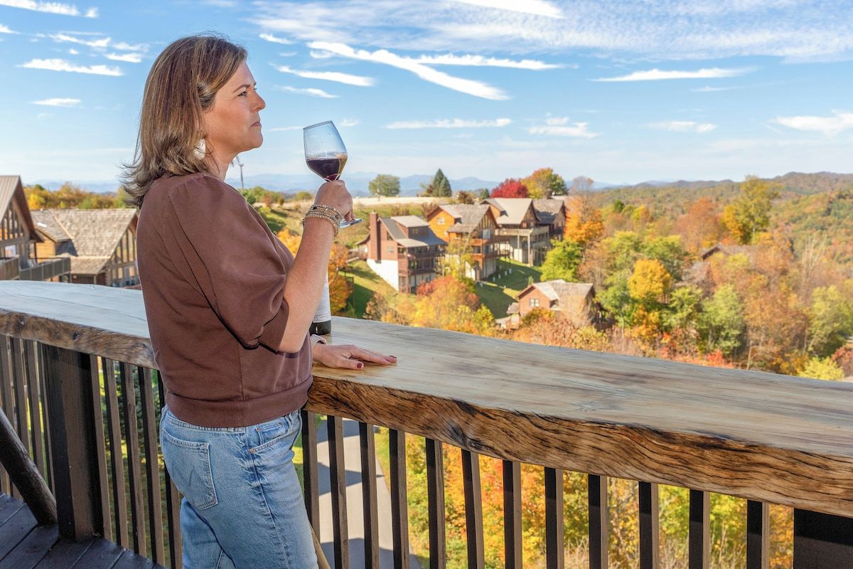Eine Person steht auf einer Holzterrasse und blickt auf ein Dorf mit Herbstlaub.