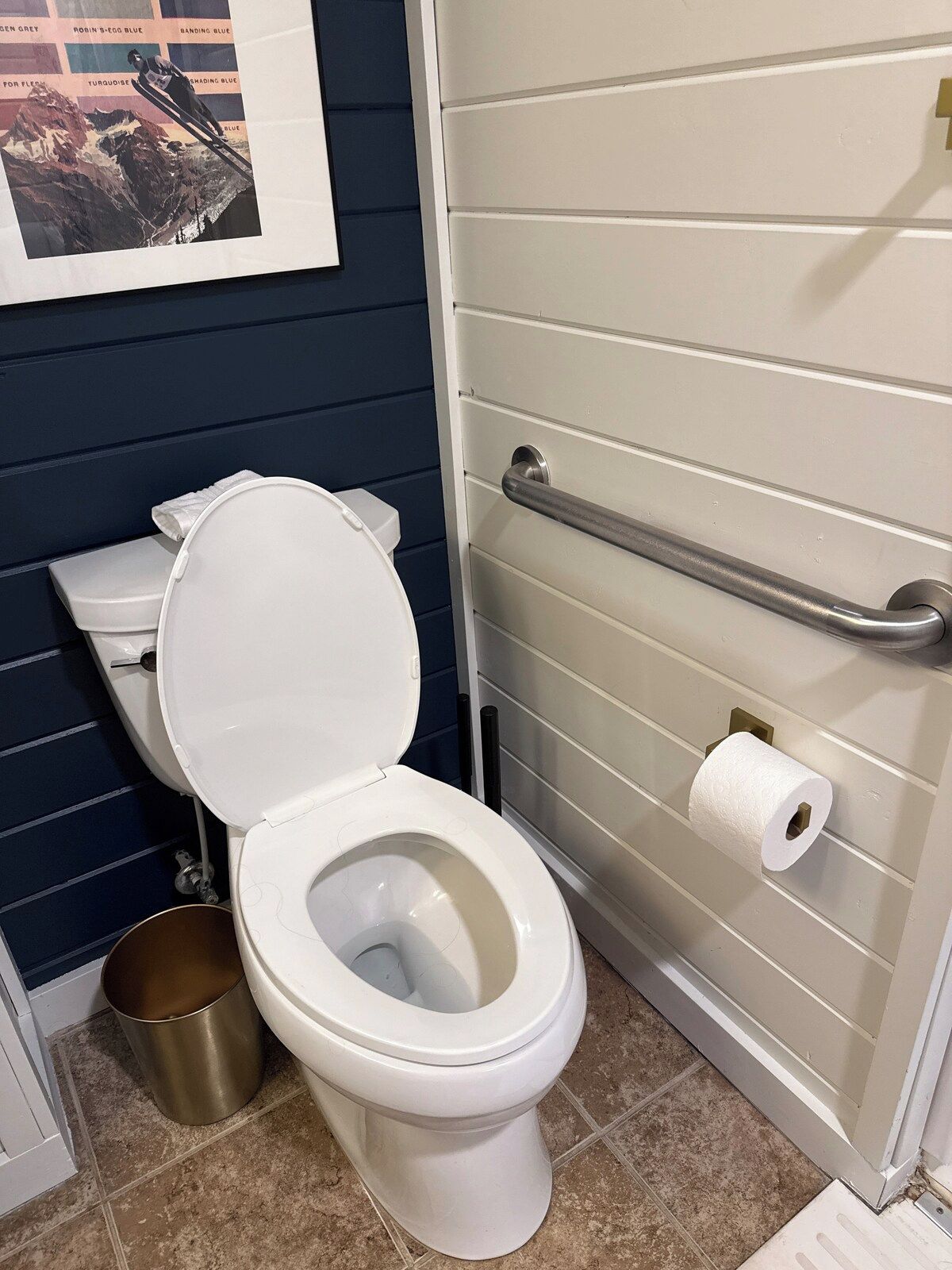 Inodoro blanco con la tapa cerrada, un bote de basura metálico y un rollo de papel higiénico en la pared.