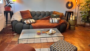 A cozy living room with a grey sofa, a glass coffee table, and decorative pillows. There are wall-mounted lights and a potted plant on a side table.