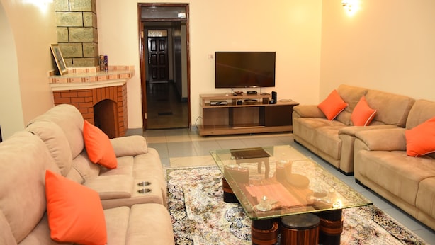 A living room with beige sofas, orange cushions, a glass coffee table, a flat-screen TV, and a brick fireplace.