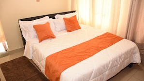 A neatly made bed with white and orange bedding, a brown rug, and a window with curtains.