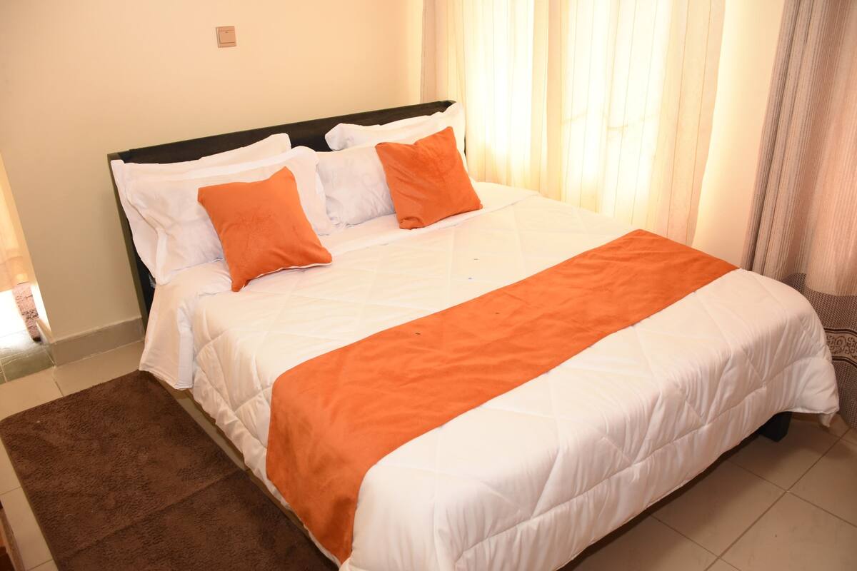 A neatly made bed with white and orange bedding, a brown rug, and a window with curtains.