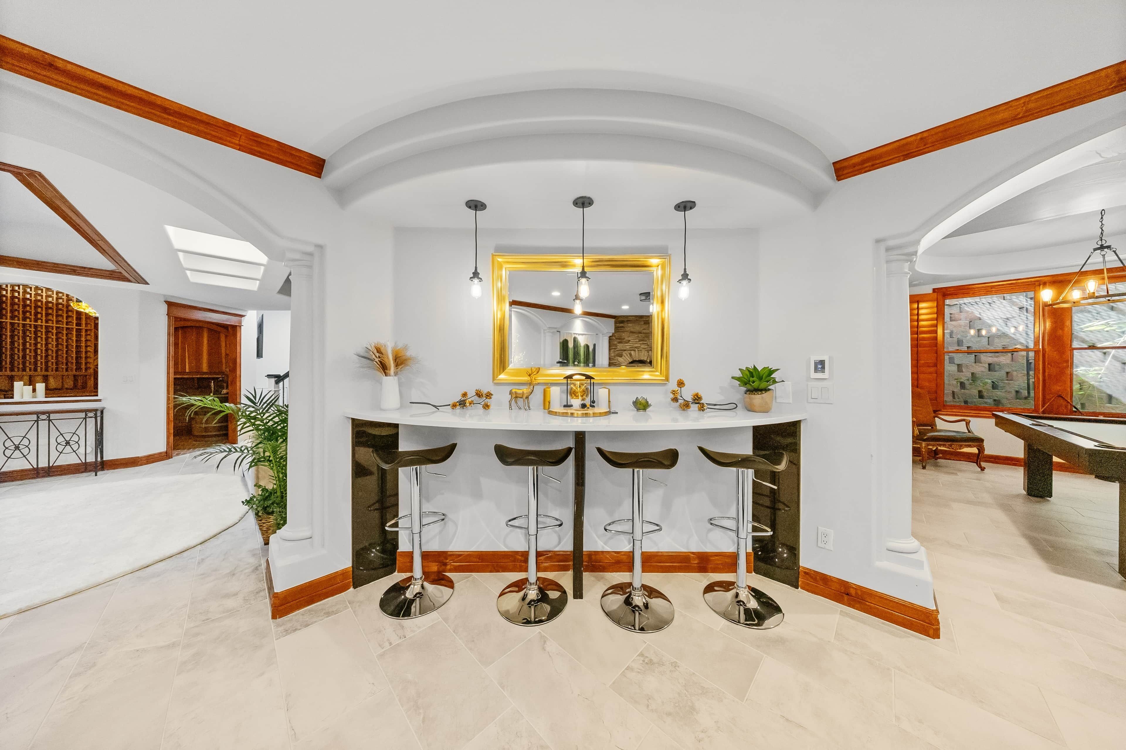 A bar area with a white countertop, three stools, and a mirror with a golden frame.