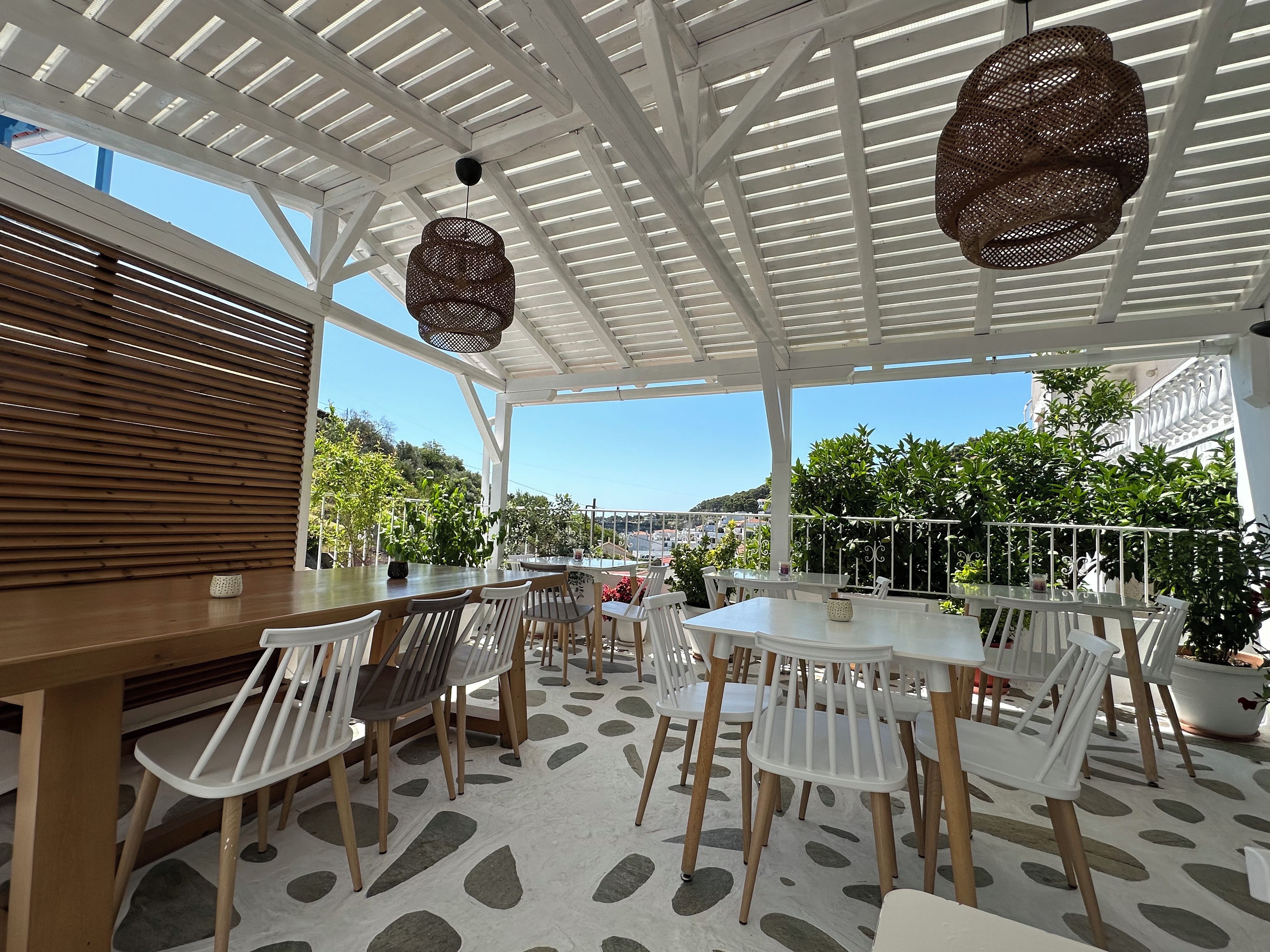 Un patio al aire libre con mesas y sillas blancas, una mesa de madera y lámparas colgantes de mimbre.