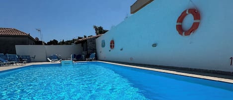 A swimming pool with clear blue water, surrounded by lounge chairs and a white wall with a lifebuoy.