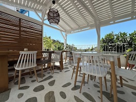 Un patio al aire libre con mobiliario blanco y de madera, suelo estampado y una pérgola blanca con una lámpara colgante.