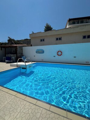 A swimming pool with clear blue water, a lifebuoy, and a stone wall with a mural. - Galini Hellenic Hospitality (Alonissos)
