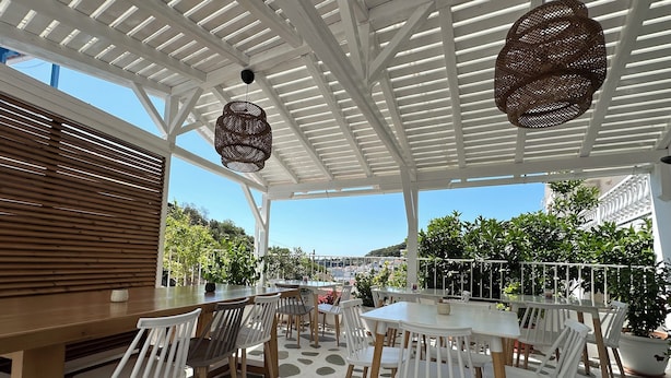 An outdoor patio with white tables and chairs, a wooden table, and hanging wicker lamps. - Galini Hellenic Hospitality (Alonissos)