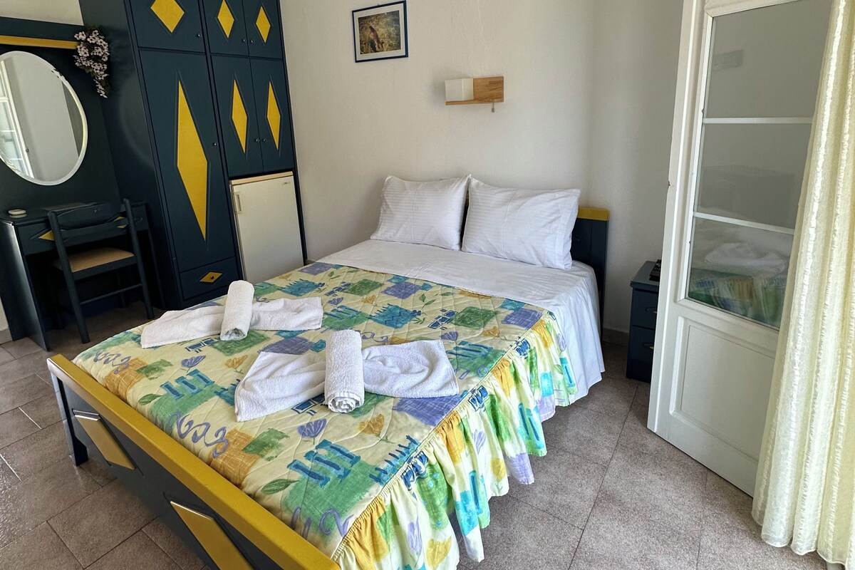 A neatly made bed with a colorful bedspread and towels, a wardrobe with geometric patterns, a small bedside table, and a window with curtains.