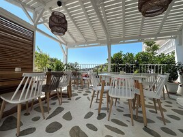 Un área de estar al aire libre techada, con mesas y sillas blancas, piso con diseño y vista a zonas verdes y edificios al fondo.