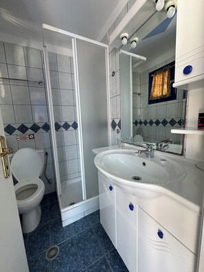 A bathroom with a white toilet, a shower enclosure, a sink with a mirror, and blue-tiled flooring. - Galini Hellenic Hospitality (Alonissos)