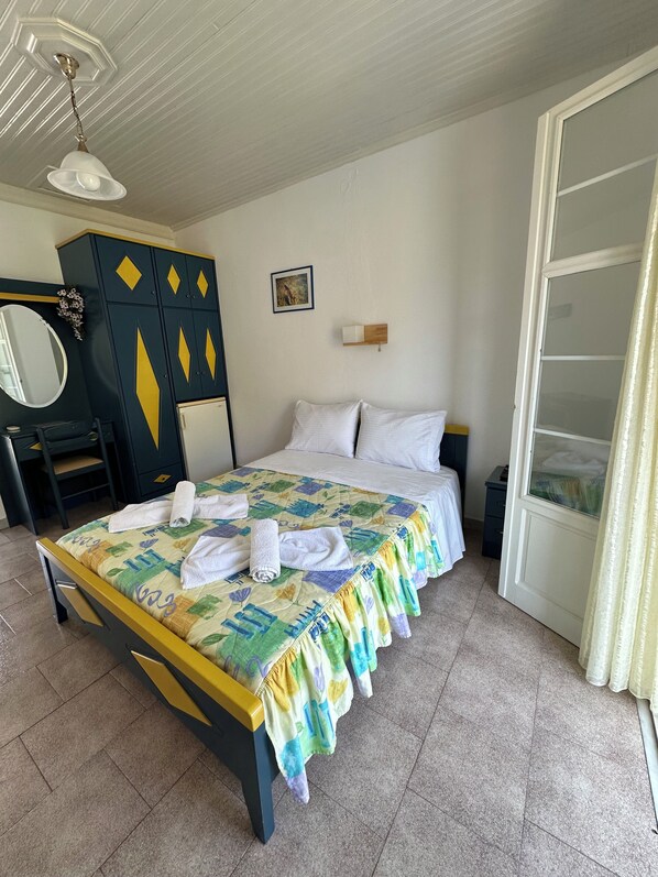A neatly made bed with a colorful bedspread and towels, a wardrobe with geometric patterns, a small bedside table, and a window with curtains. - Galini Hellenic Hospitality (Alonissos)