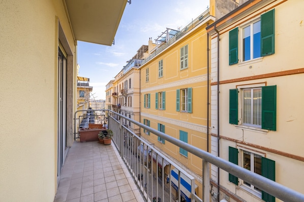 A narrow balcony with a metal railing overlooking a row of yellow buildings with green shutters.