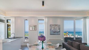 Un comedor moderno con una mesa de madera dispuesta para seis personas, ventanas amplias con vista al mar y un rincón de estar acogedor con un sofá.