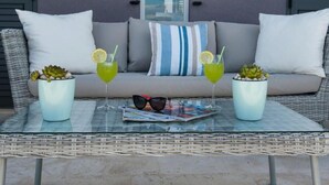 Un sofá de mimbre con cojines, dos suculentas en macetas, una bandeja con gafas de sol y una revista, y dos vasos de bebida verde con rodajas de limón y pajitas.