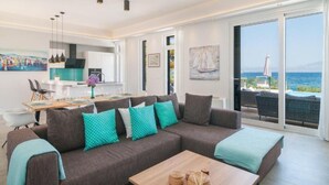 Una sala de estar moderna con sofá, mesa de centro y obras de arte. Hay una zona de cocina con una mesa de comedor y sillas. La habitación tiene ventanas amplias con vistas al mar.