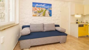 A cozy living room with a sofa, a painting of a coastal town, and a kitchen area. - Studio with air conditioning and barbecue facilities (Crikvenica)