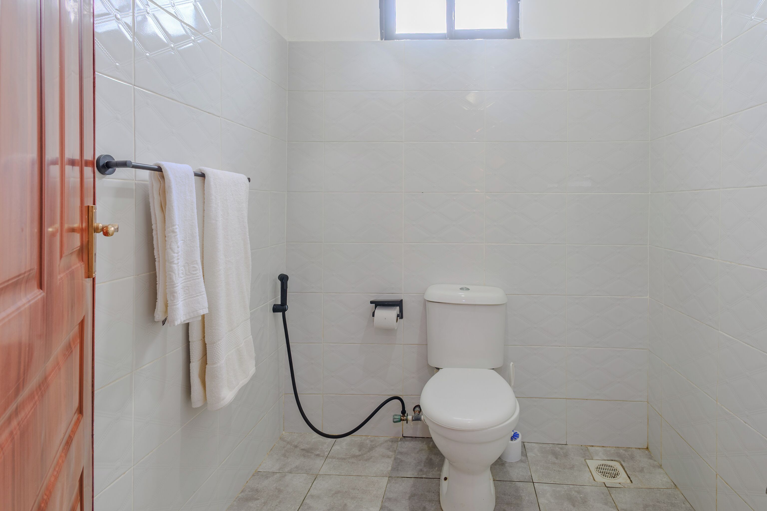 A small, clean bathroom with white tiles, a toilet, a towel rack with two towels, and a window above the toilet.