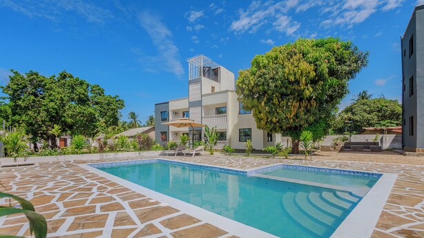 Una piscina con un diseño geométrico de azulejos, rodeada de vegetación y un edificio moderno con balcones.