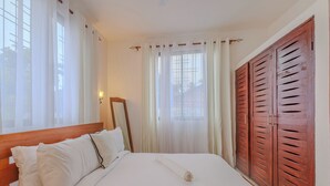 Un dormitorio con cabecera de madera, una cama grande con ropa blanca, un armario de madera y cortinas traslúcidas en las ventanas.