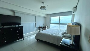 A modern hotel room with a bed, a TV on a dresser, a desk, and a window with a view of the sea. - Ocean Cafe I Spacious 1 BDR Apt With Sea View (Panama City)
