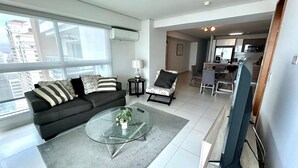 A modern living room with a glass coffee table, a sofa with striped cushions, a side table with a lamp, and a view of the city through large windows. - Ocean Cafe I Spacious 1 BDR Apt With Sea View (Panama City)