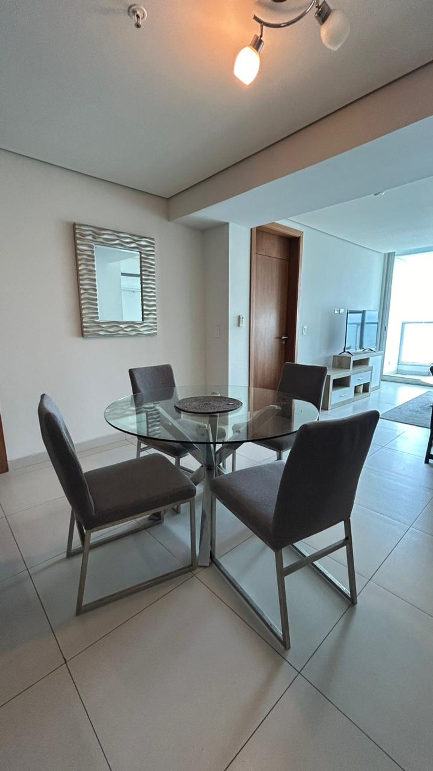 A modern dining area with a glass table and four chairs, a decorative mirror, and a wooden door leading to another room.