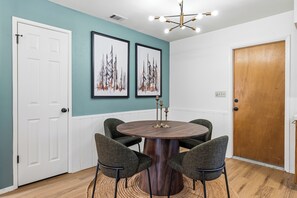 A dining area with a round wooden table, four upholstered chairs, teal walls, two framed artworks, a wooden door, and a ceiling light fixture.