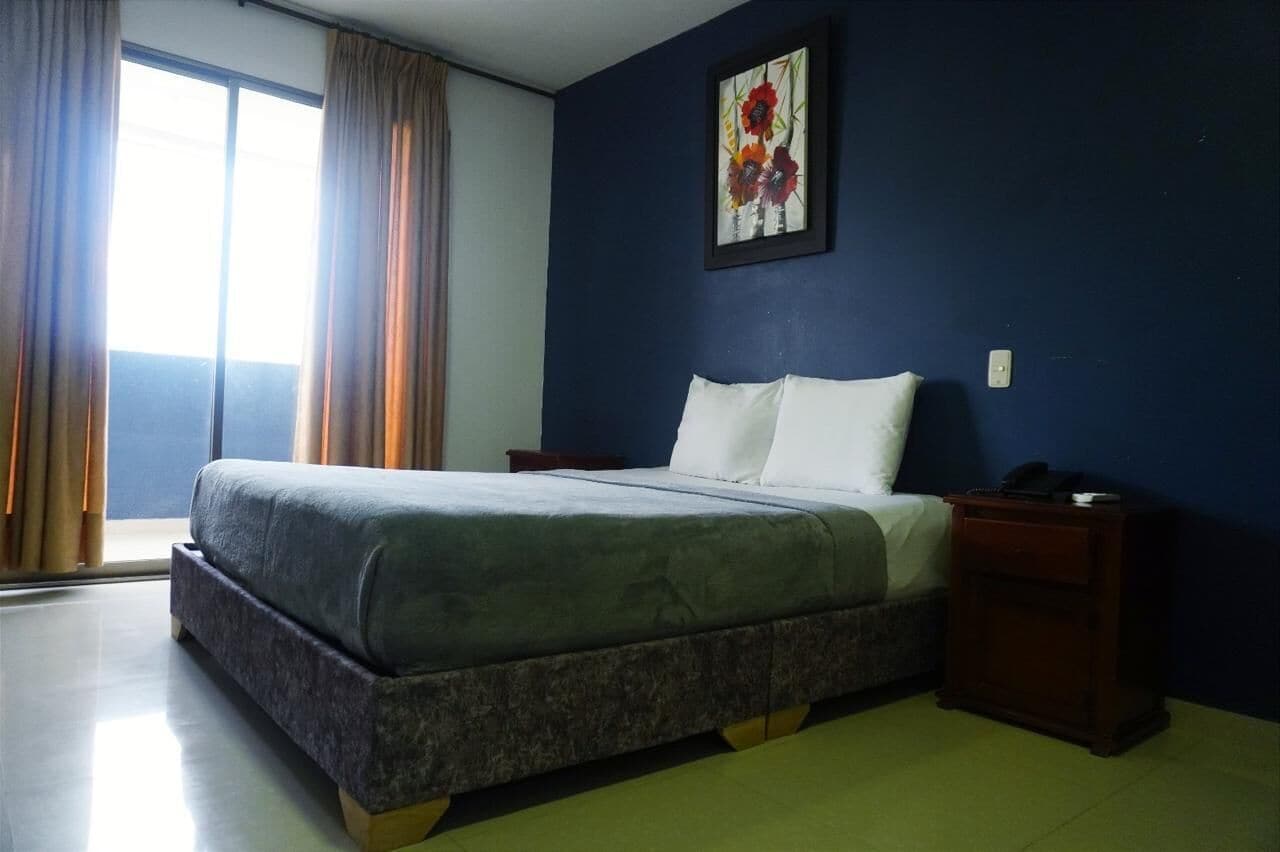 A bedroom with a bed, a nightstand, a painting on the wall, and a window with curtains.