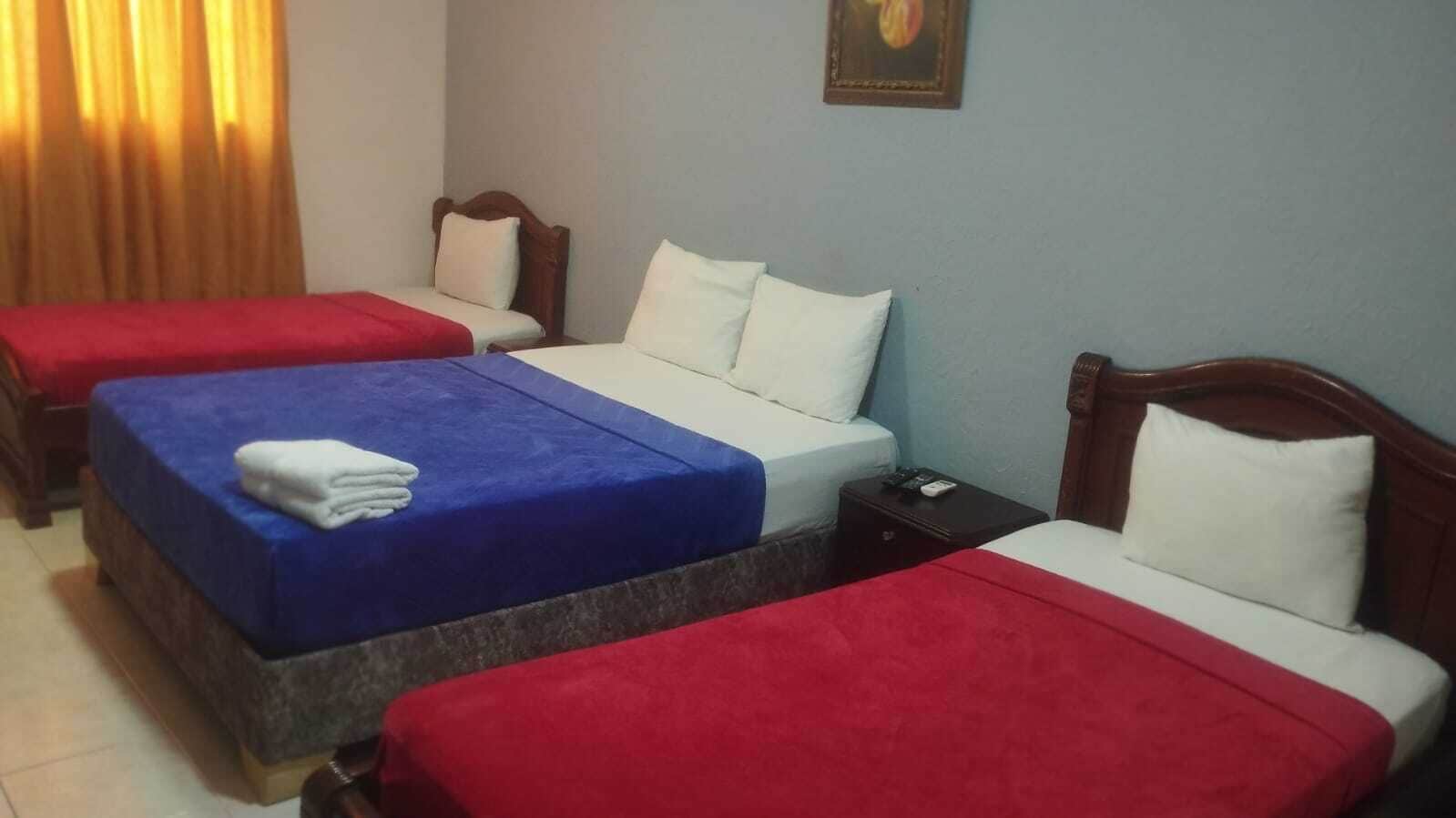 A hotel room with three single beds, each with a red and white bedspread. There is a wooden nightstand between the beds, and a small table with a remote control on it.
