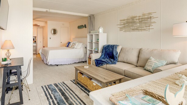A well-lit, spacious room with a bed, a sofa, a coffee table, and a desk with a lamp. There is a wall-mounted shelf with decorative items and a wall art piece. - 1 Block to Beach! Ocean City Condo w/ Pool Access (Ocean City)