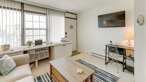 A modern living room with a sofa, a coffee table, a desk with a chair, a TV mounted on the wall, and large windows with blinds. - 1 Block to Beach! Ocean City Condo w/ Pool Access (Ocean City)