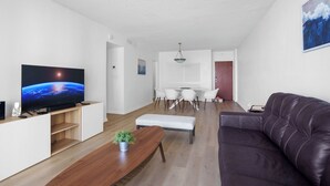 A modern living room with a flat-screen TV, a dark leather sofa, a wooden coffee table, and a dining area with white chairs and a table. - Condo w/2 Queen beds and Ocean View | Tides (Hollywood)