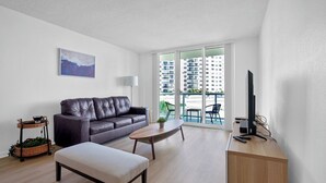 A modern living room with a leather sofa, a wooden coffee table, a small side table with a plant, and a television on a wooden cabinet. - Condo w/2 Queen beds and Ocean View | Tides (Hollywood)