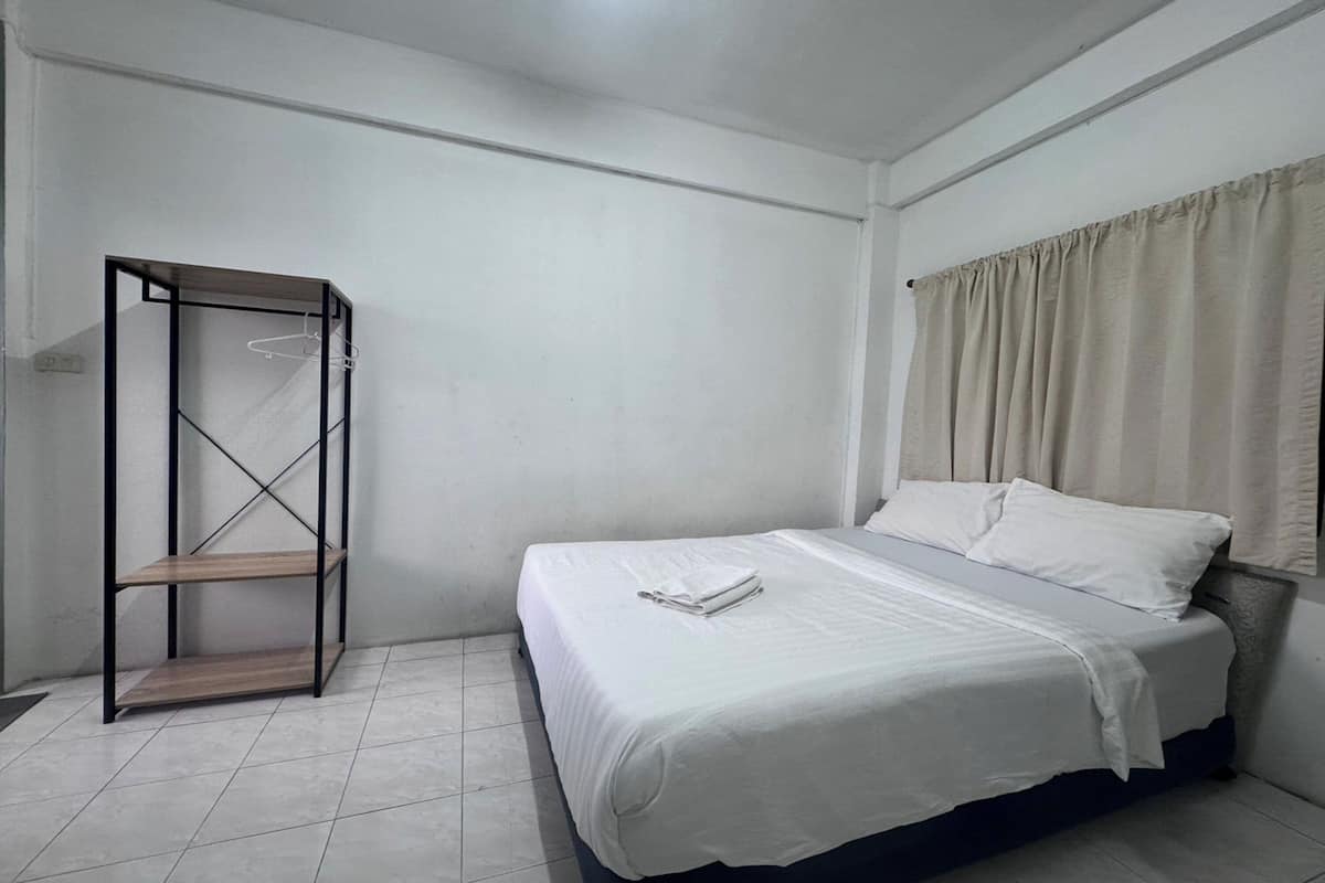 Una cama individual con colchón y almohadas blancas, un estante de madera y una cortina beige en la ventana.