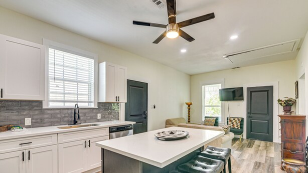 A modern kitchen with white cabinets, a central island with bar stools, a ceiling fan, and a large window with blinds. - Close to Beach: Walkable Galveston Getaway! (Galveston)