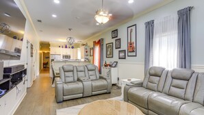 A spacious living room with a gray leather sofa, a small white side table, and a woven ottoman. A kitchen with white cabinets and a dining area with a table and chairs is visible in the background. - Family Home w/ Fenced Backyard in New Orleans! (New Orleans)