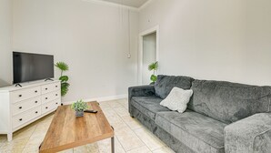 Ein Wohnzimmer mit Flachbildfernseher auf einer weißen Kommode, einem grauen Sofa mit weißem Kissen, einem Holztisch und zwei Blumentöpfen.