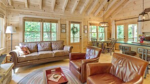 A wooden cabin interior with a sofa, chairs, a dining table, and a kitchen area. - 4 Mi to Fall Branch Falls: Peaceful Cabin w/ Deck (Cherry Log)