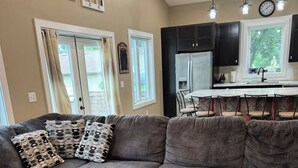 A modern living room with a sectional sofa, ottoman, and a kitchen area with dark cabinets and a window. - Cozy, beautiful, family friendly, 3-bedroom, newly built cabin on Lake Latoka (Alexandria)