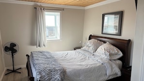 Un dormitorio con techo de madera, cama con ropa de cama estampada, un ventilador de pie, una ventana con cortinas y un cuadro en la pared.
