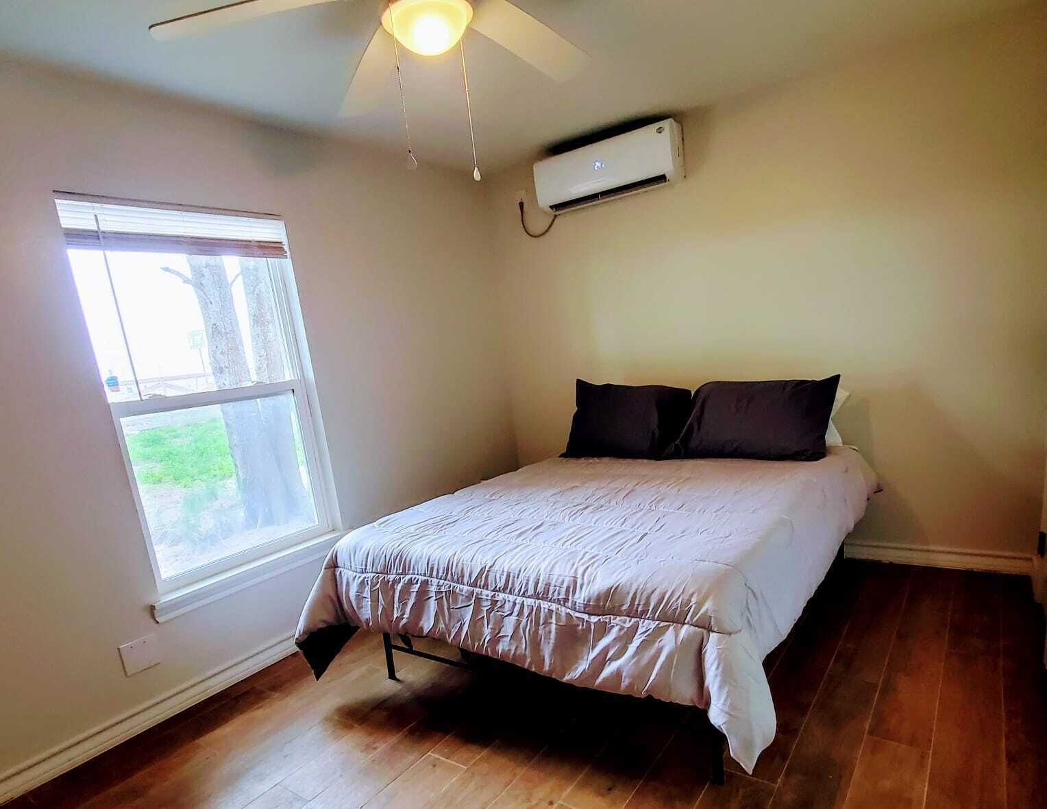 A bedroom with a bed, a ceiling fan, an air conditioning unit, and a window with a view of greenery.