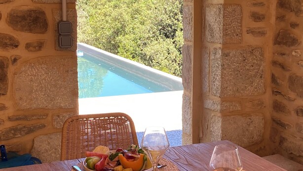 A stone-walled room with a wooden table set for a meal, overlooking a pool and lush greenery.