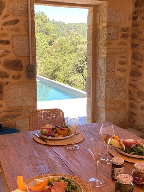 A stone-walled room with a wooden table set for a meal, overlooking a pool and lush greenery.