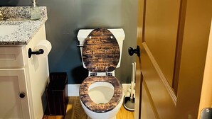 A bathroom with a wooden toilet seat, a sink with a granite countertop, a wall-mounted light, and a door with a silver handle. - Bourbon Under the Stars Drive-In-Puzzle Room Experience (Frankfort)