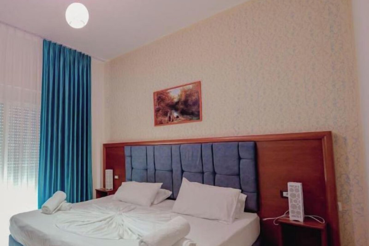 A hotel room with a large bed, a headboard, a nightstand, a painting on the wall, and a window with curtains.