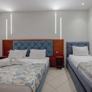 A hotel room with two beds, each with a wooden headboard and a blue upholstered headboard. There are white pillows and a heart-shaped towel on the left bed. The room has a tiled floor and a plain white wall with a modern light fixture.