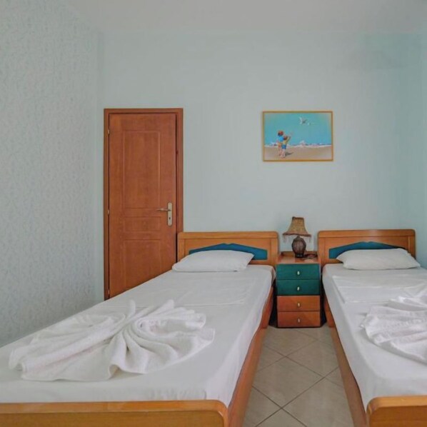A hotel room with two beds, a wooden door, a painting on the wall, and a nightstand with a lamp. - Hotel Divo Palace (Ksamil)